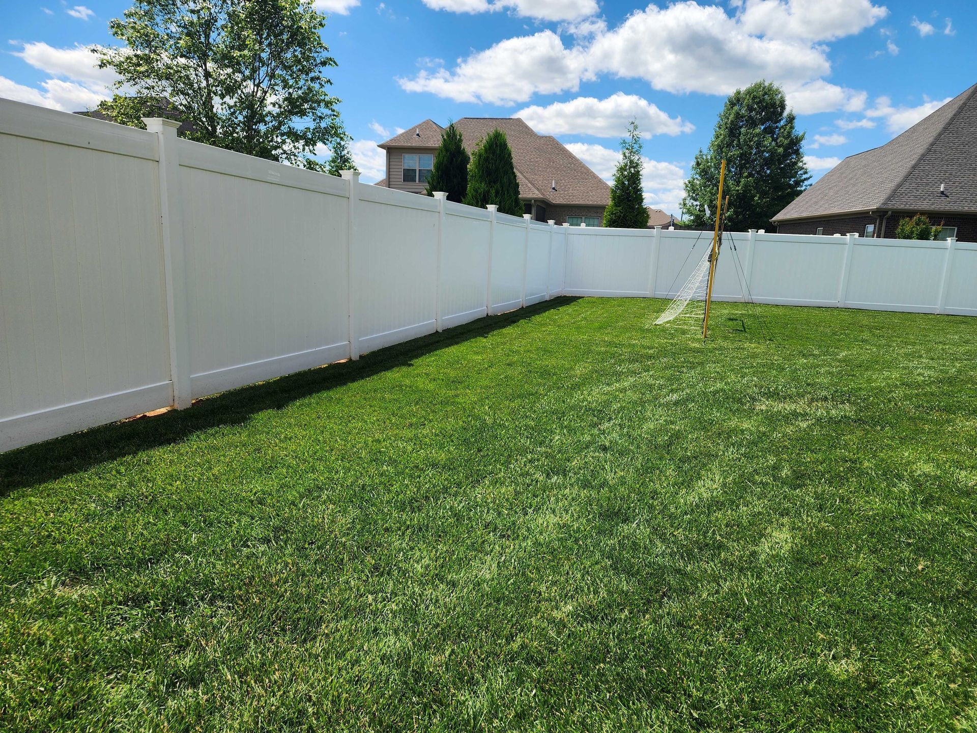 White vinyl fence encloses a green grassy backyard on a sunny day; houses visible in the distance.