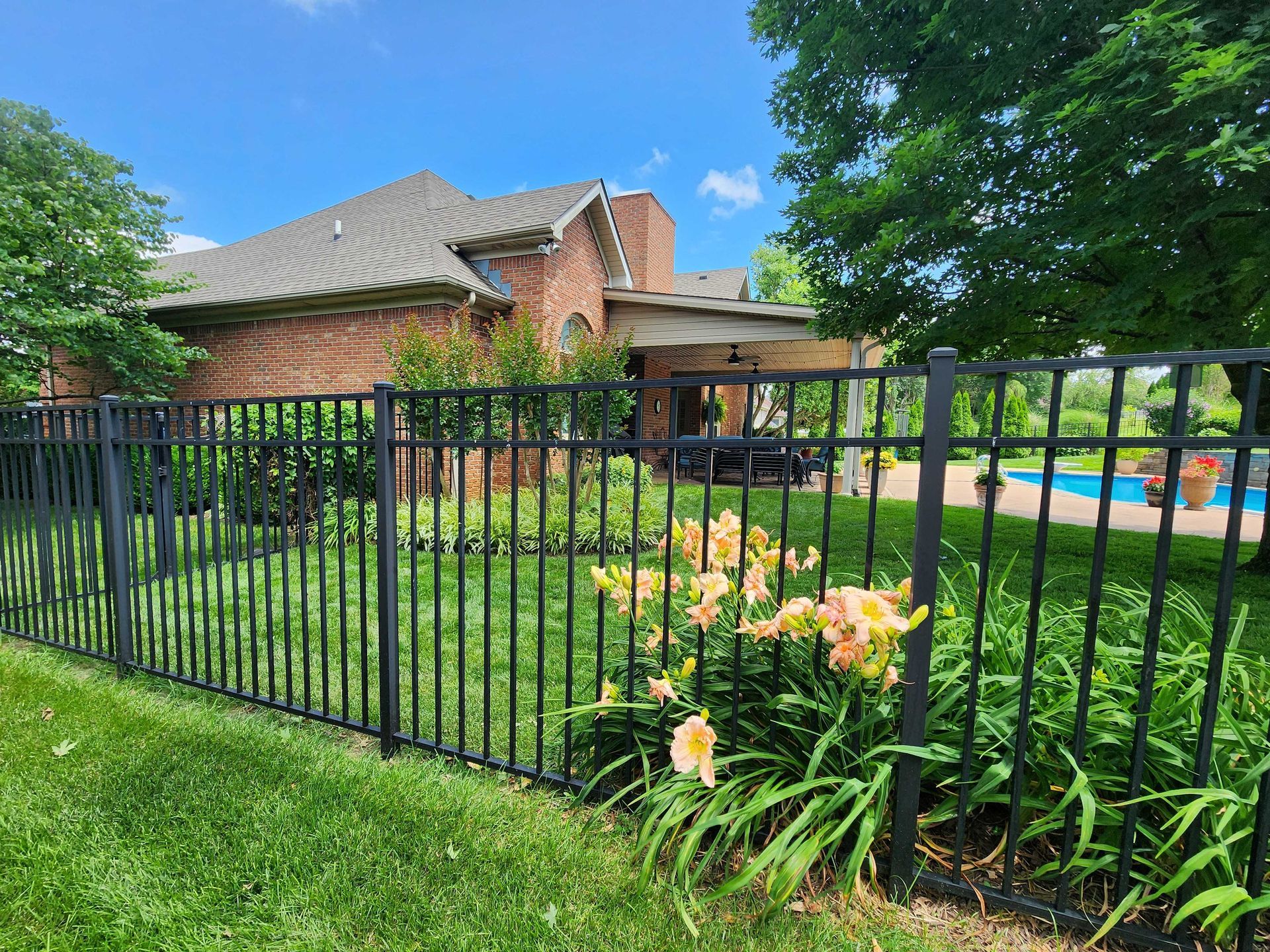 Black metal fence surrounding a yard with a brick house, flowers, and a pool on a sunny day.