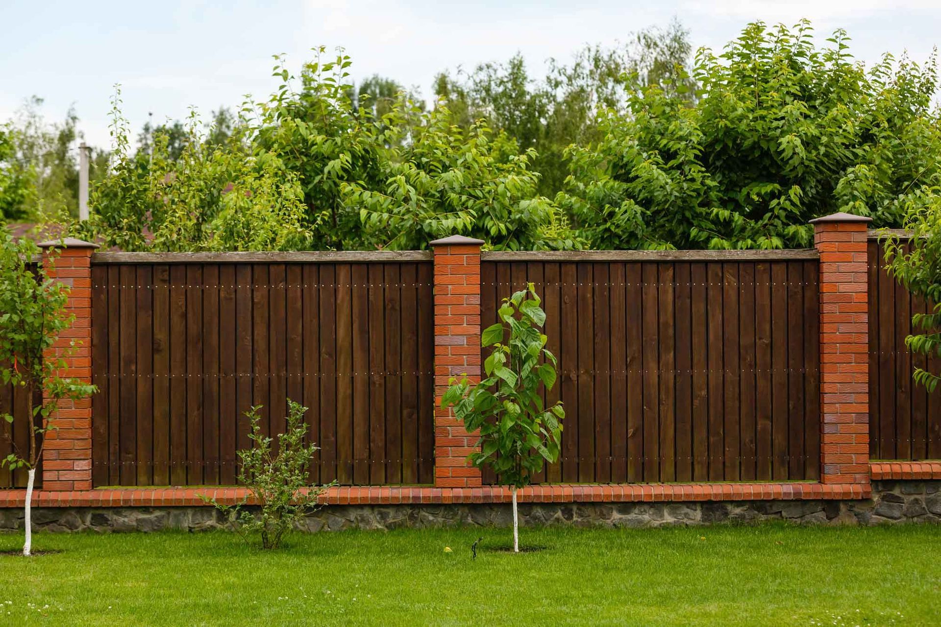 Wooden fence with brick pillars borders a green lawn and leafy garden. Wooden fence with brick pillars borders a green lawn and leafy garden.