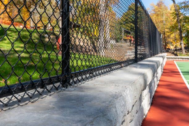 A chain link fence surrounds a tennis court in a park.