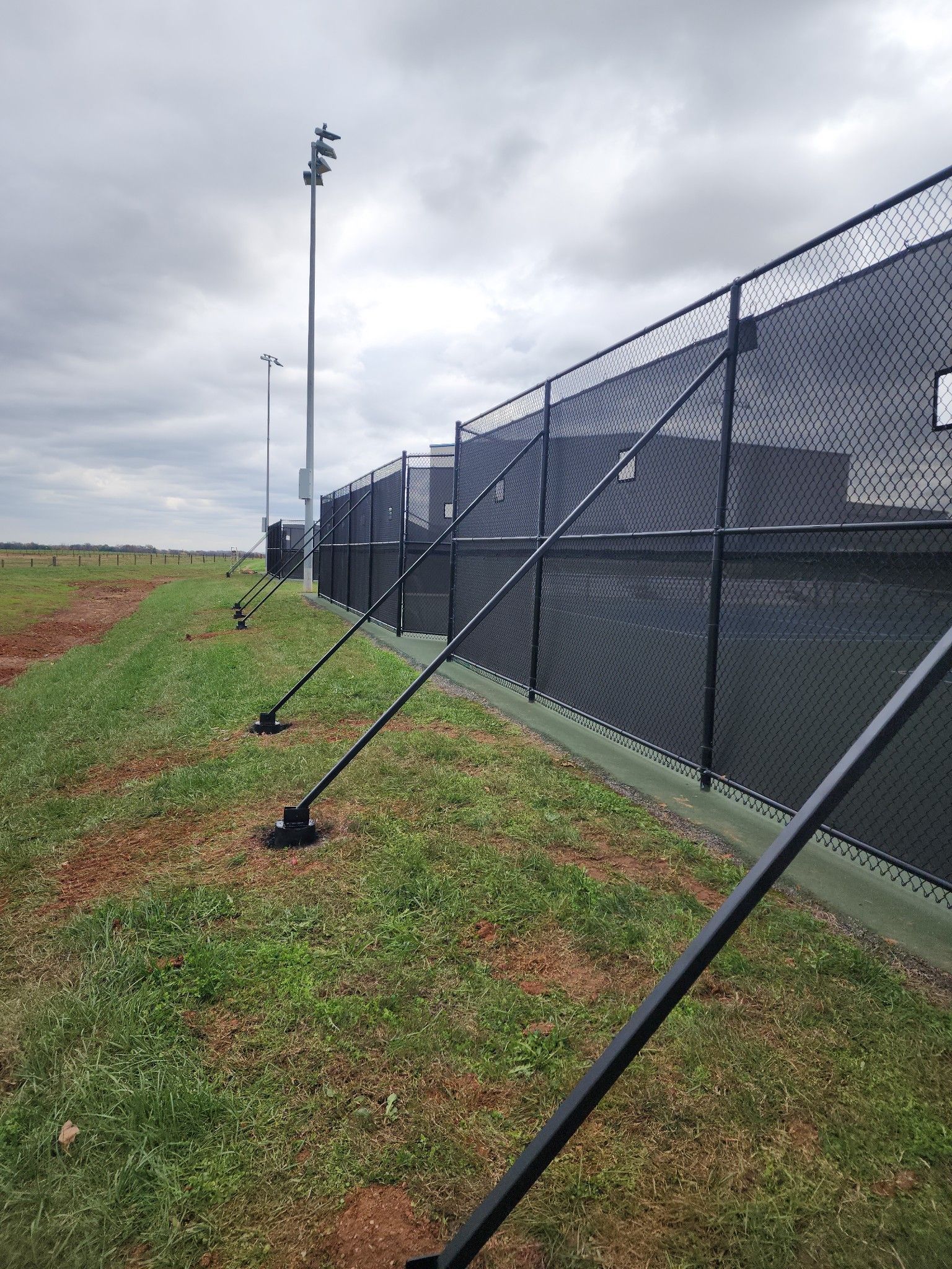 Black chain link fence along a grass-covered earthen berm, under a cloudy sky.