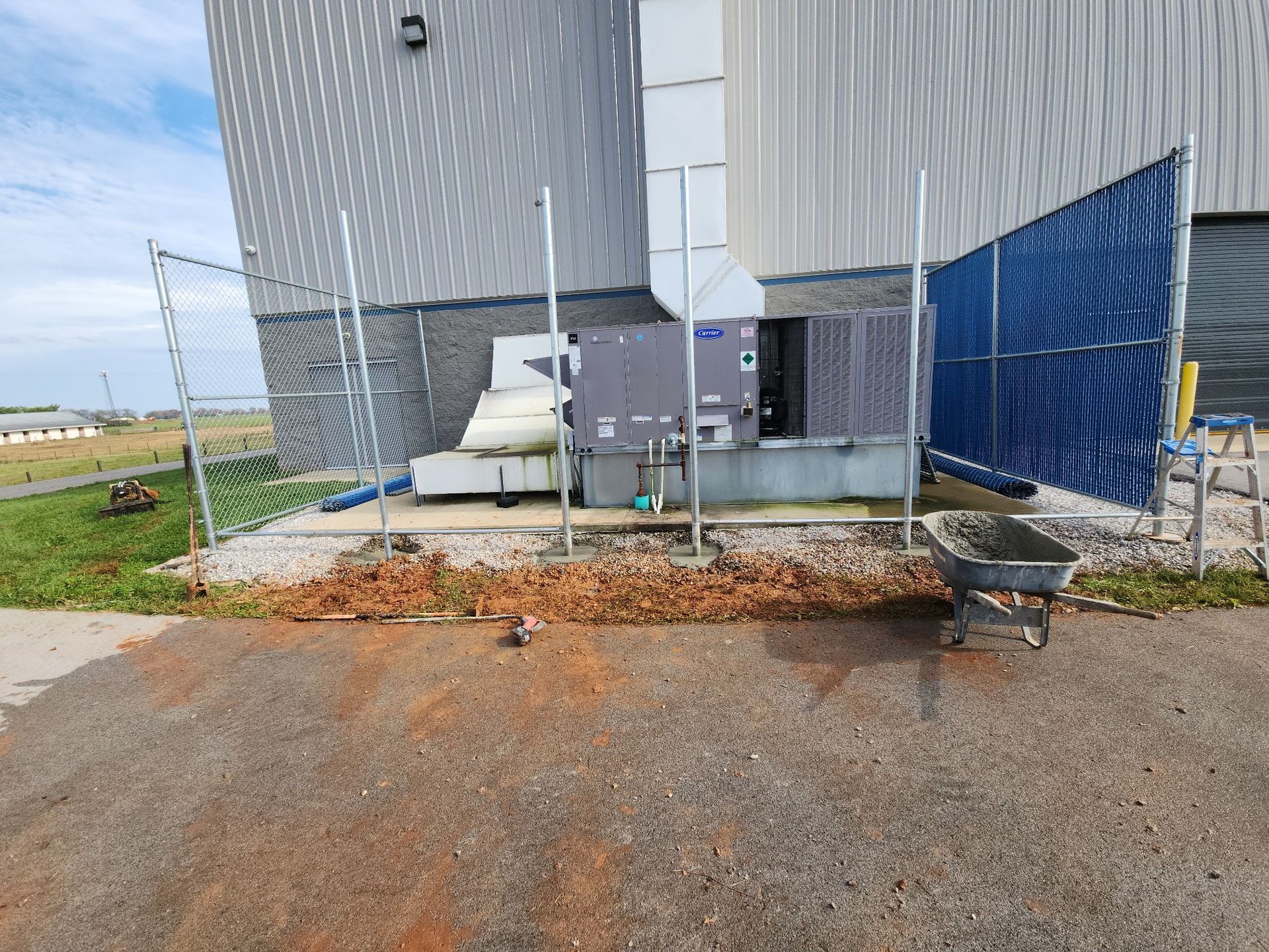 Outdoor HVAC unit next to building, protected by fencing; ground covered in mulch and dirt.
