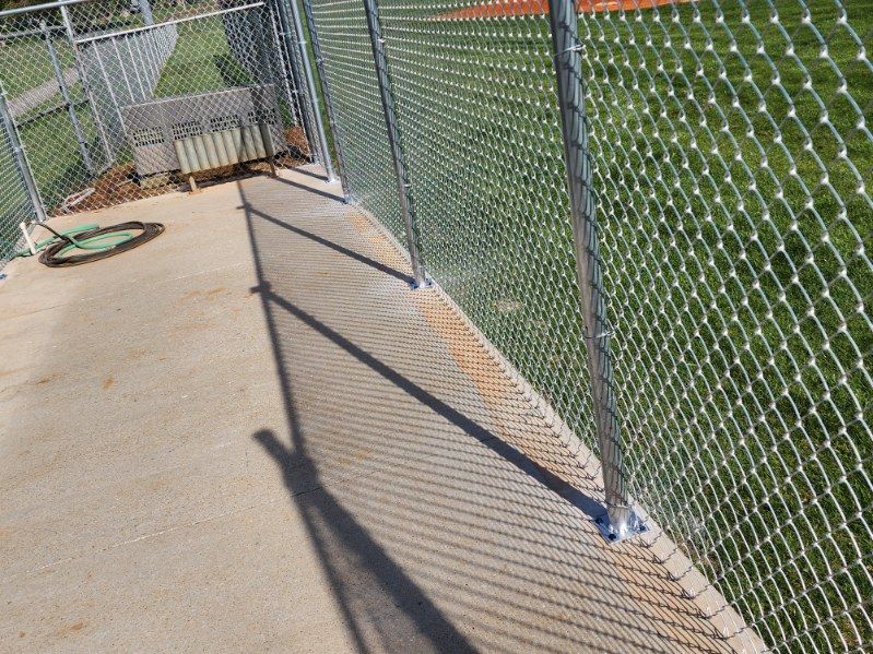 A chain-link fence atop a concrete barrier casts a shadow on the ground, near a baseball field.