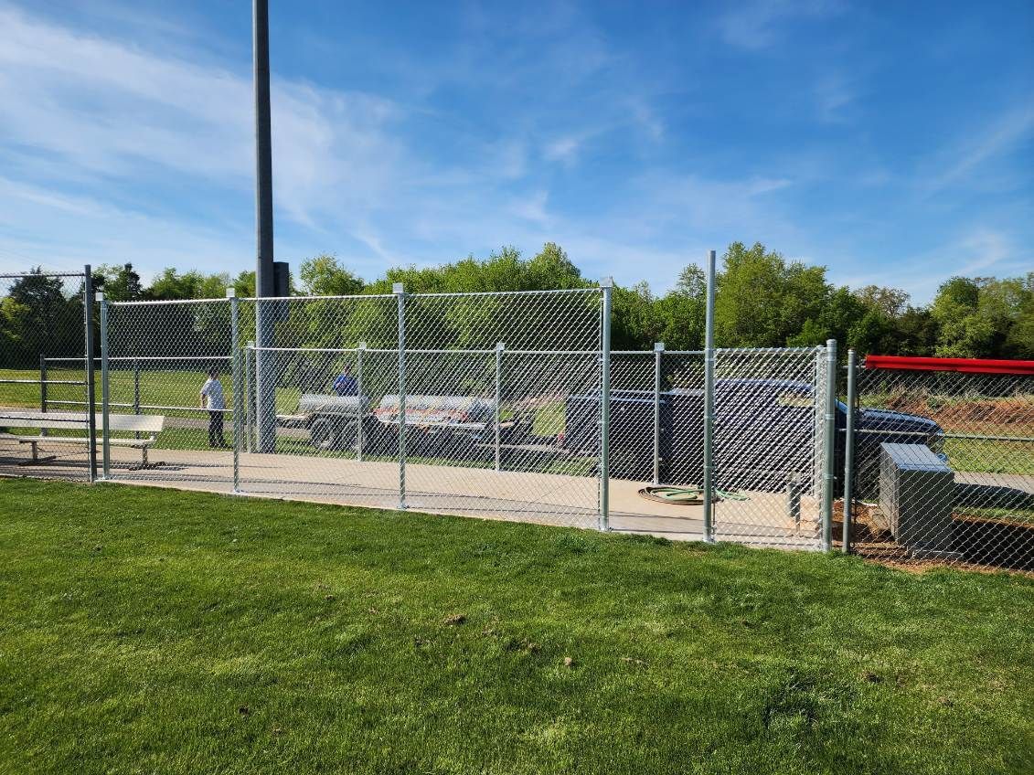 Chain-link fence surrounds an equipment area at a baseball field, grass in foreground, blue sky above.
