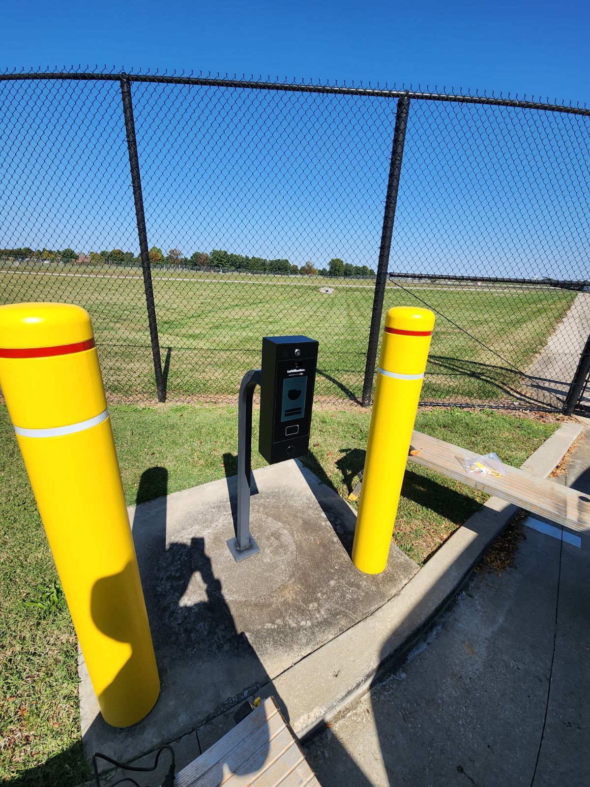 Yellow bollards flank a black access control box with a fence and field in the background.