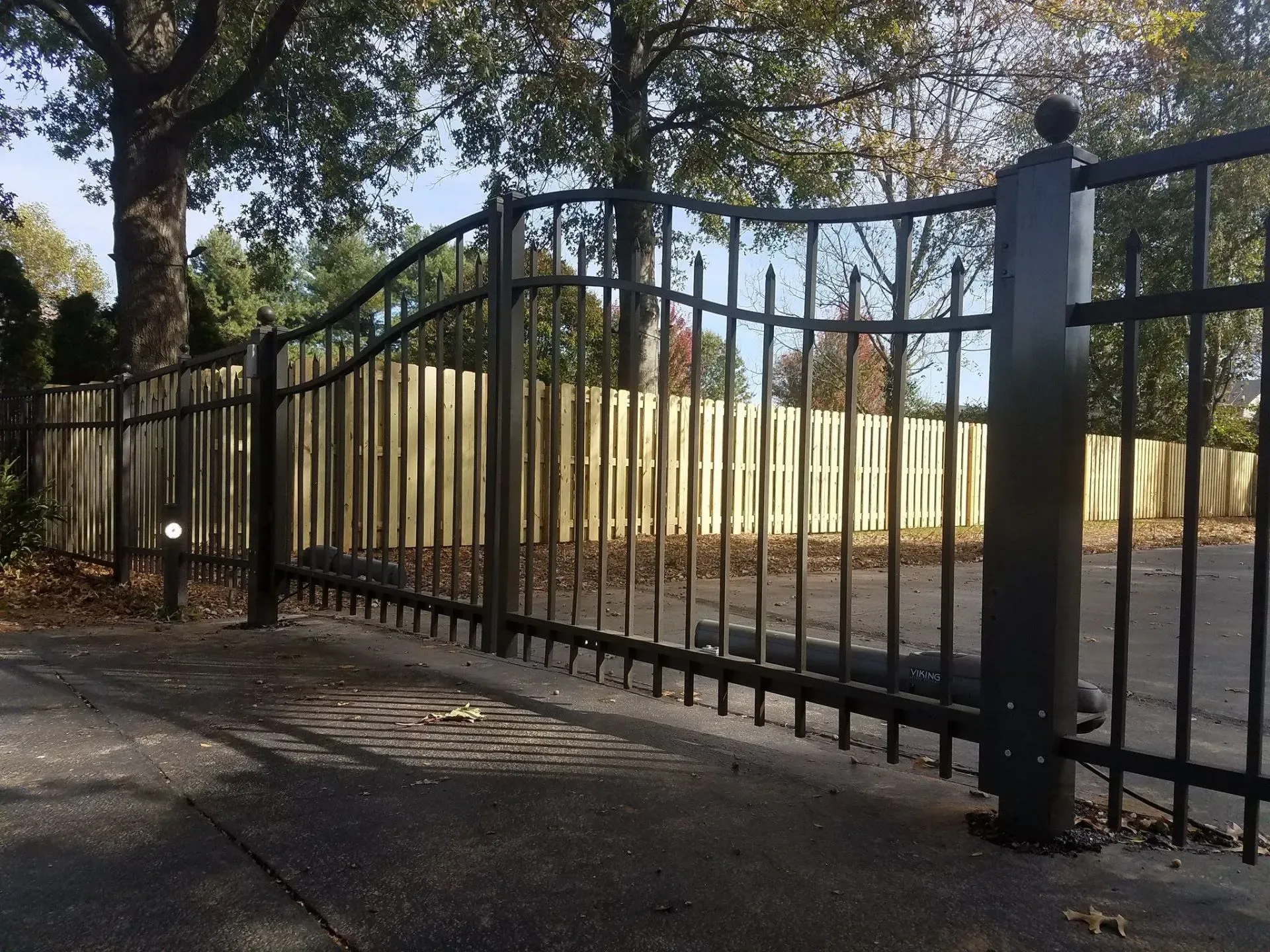Black metal gate and fence with wooden fence in the background, trees overhead.