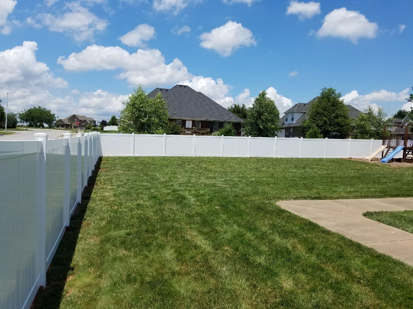 White vinyl fence encloses a grassy backyard, houses in the background, blue sky with clouds.