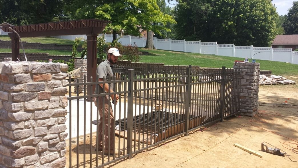 Man installs iron gate between stone pillars in a yard. White fence and trees in background.