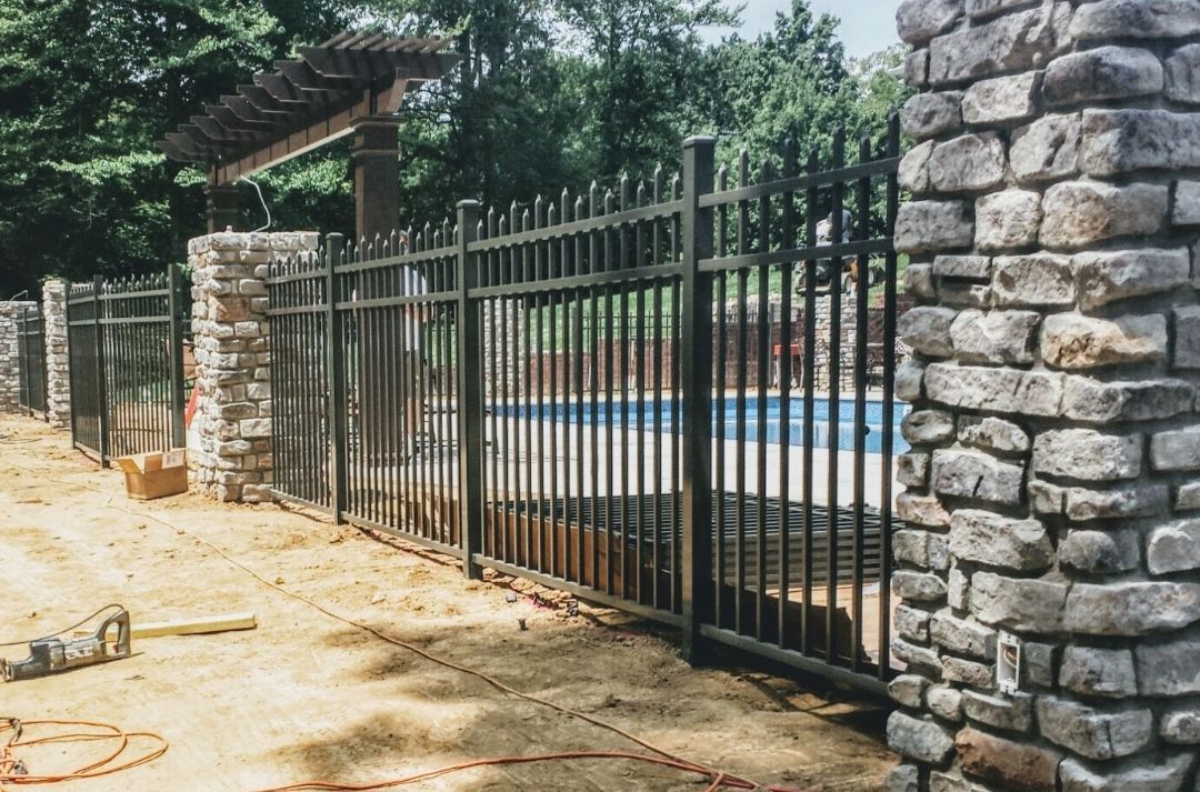 Black metal fence around a pool, flanked by stone columns. Brown dirt in foreground, trees in background.