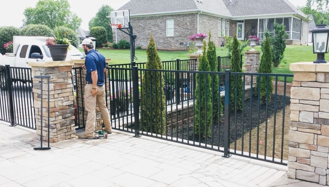 Man standing near a black metal fence with stone pillars and landscaping in front of a house.