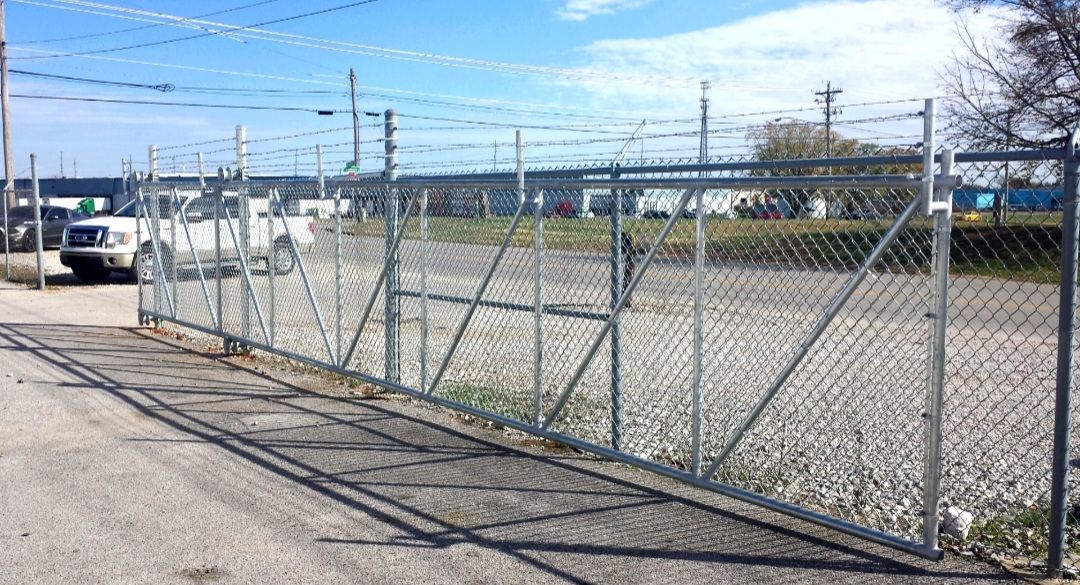 Chain-link fence with a sliding gate, topped with barbed wire, in a parking lot. A white van is parked nearby.