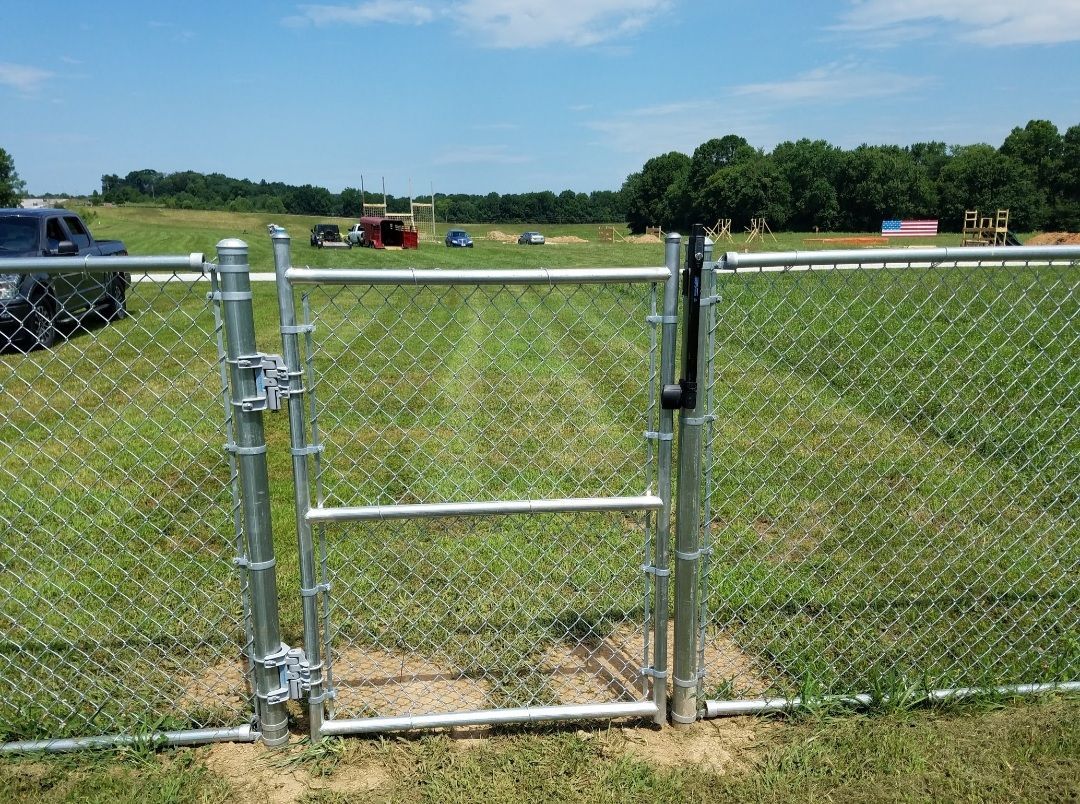 Chain-link fence gate open into a grassy field, with trees in the background under a blue sky.
