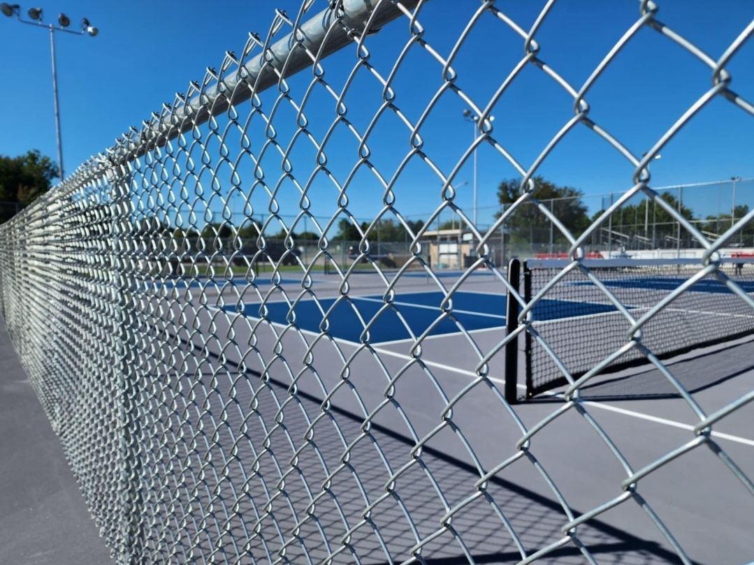 Chain-link fence in foreground, blue tennis court with net visible through the fence on a sunny day.