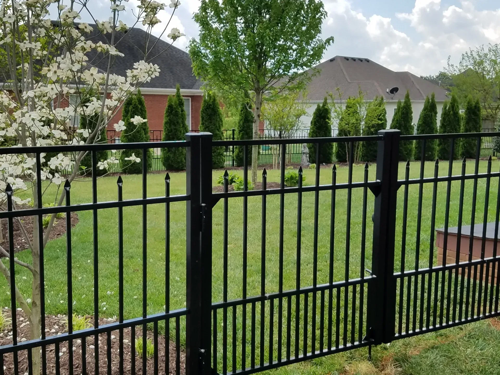 A black fence surrounds a lush green yard with a house in the background