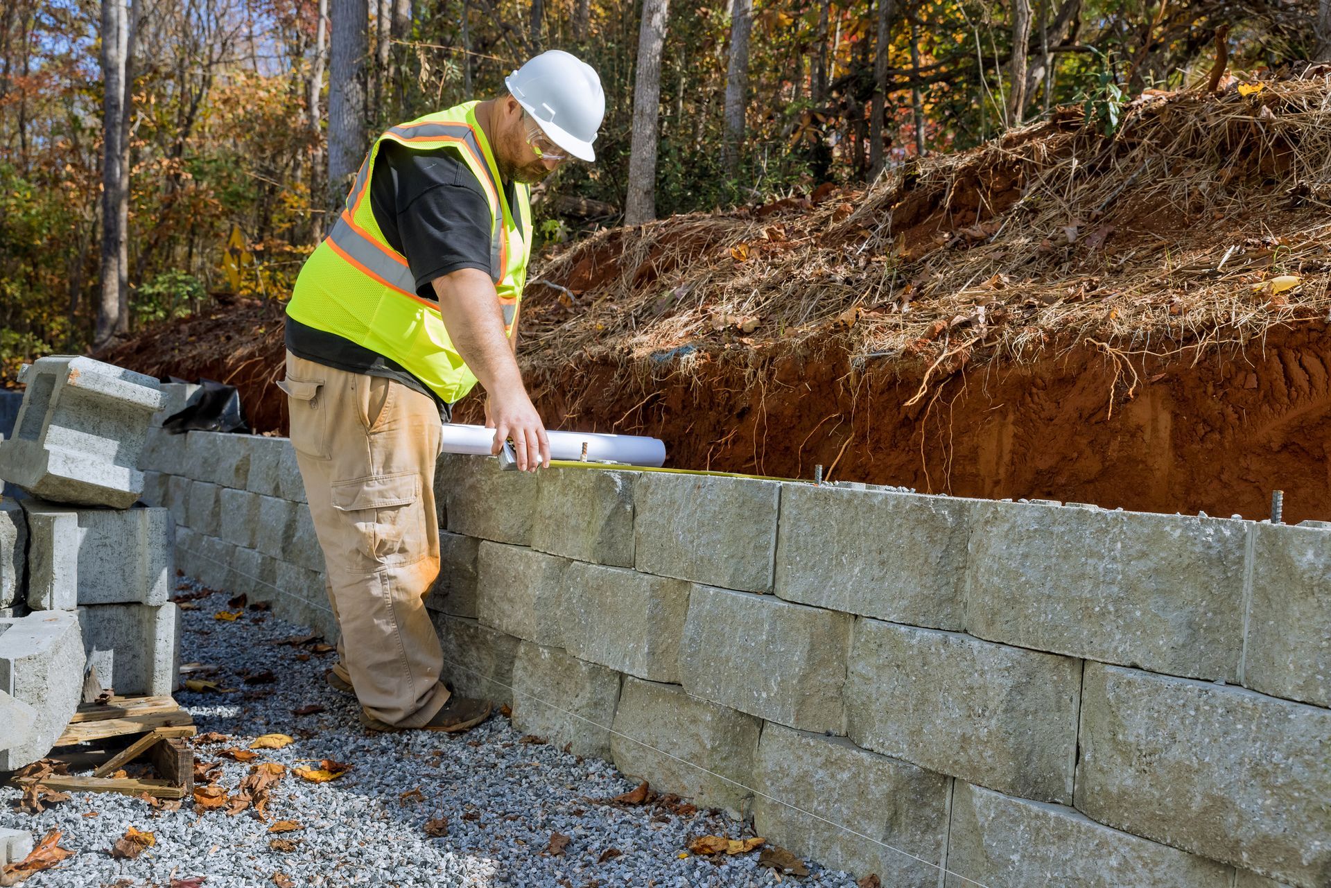 Retaining wall construction worker reviewing plans on concrete block wall at wooded job site.