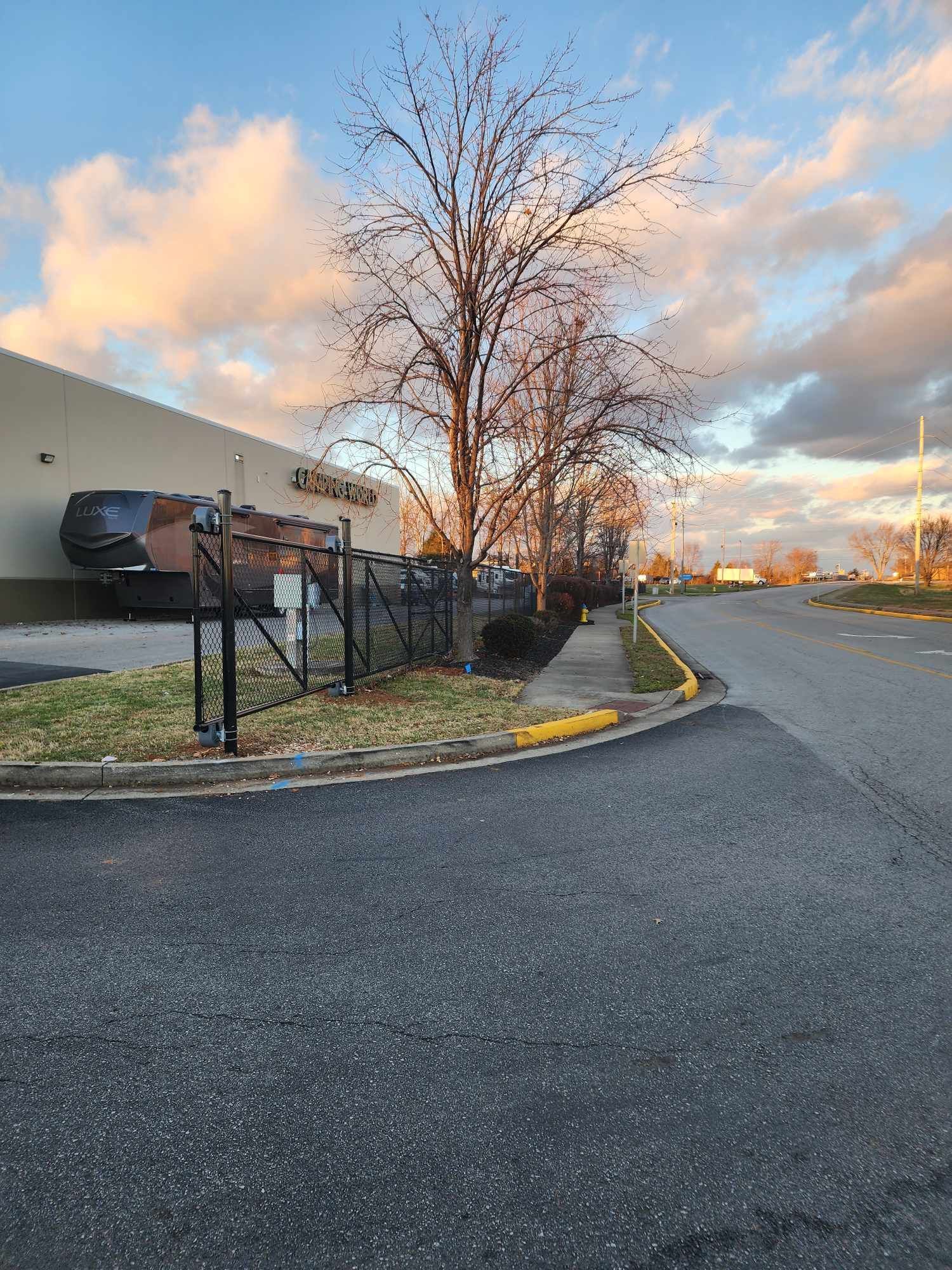 Paved road curves near building with loading dock; a tree and fence are visible, with a cloudy sky overhead.