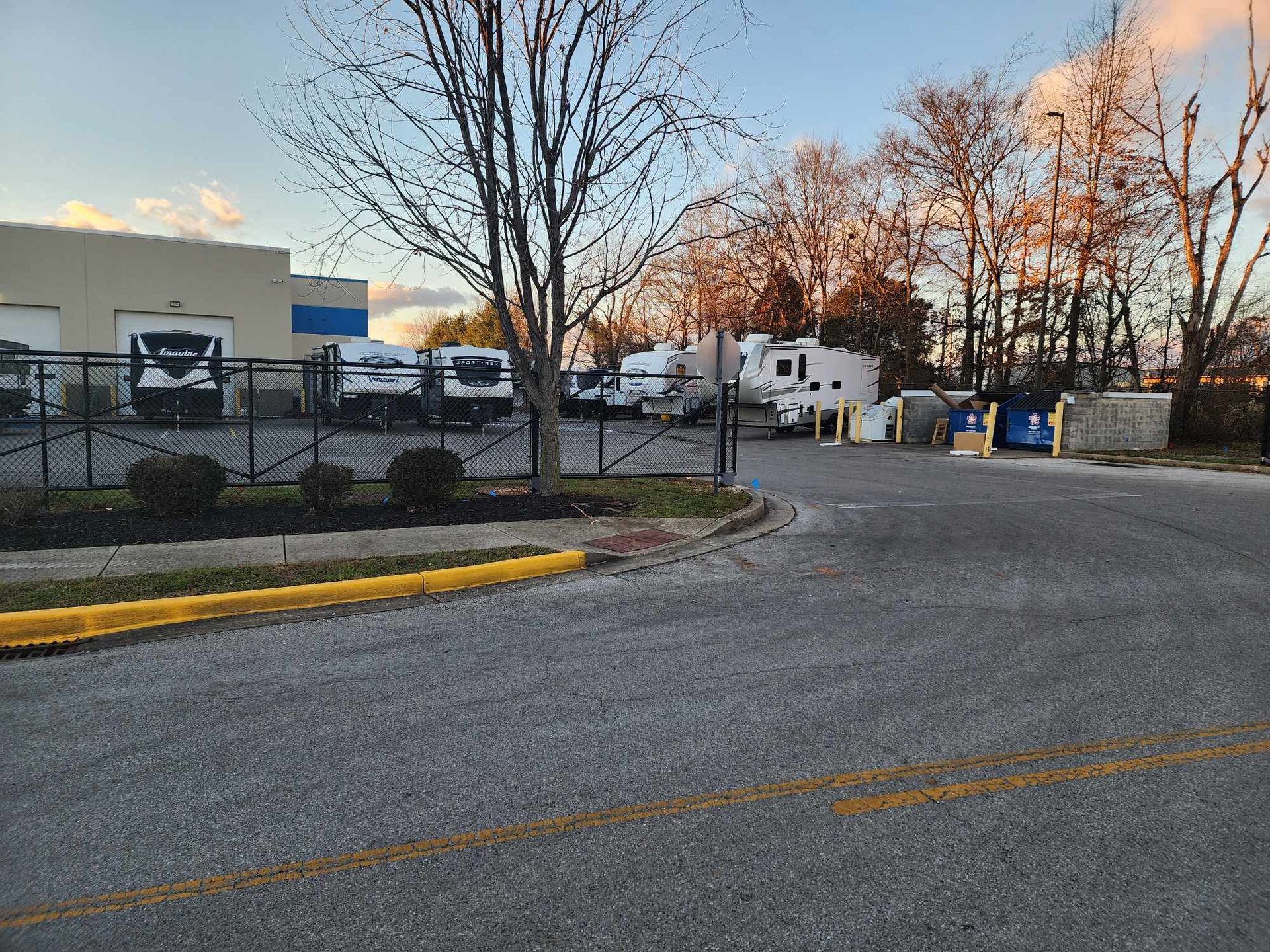 A paved lot with RVs and trucks, a metal fence, and a building under a partly cloudy sky.