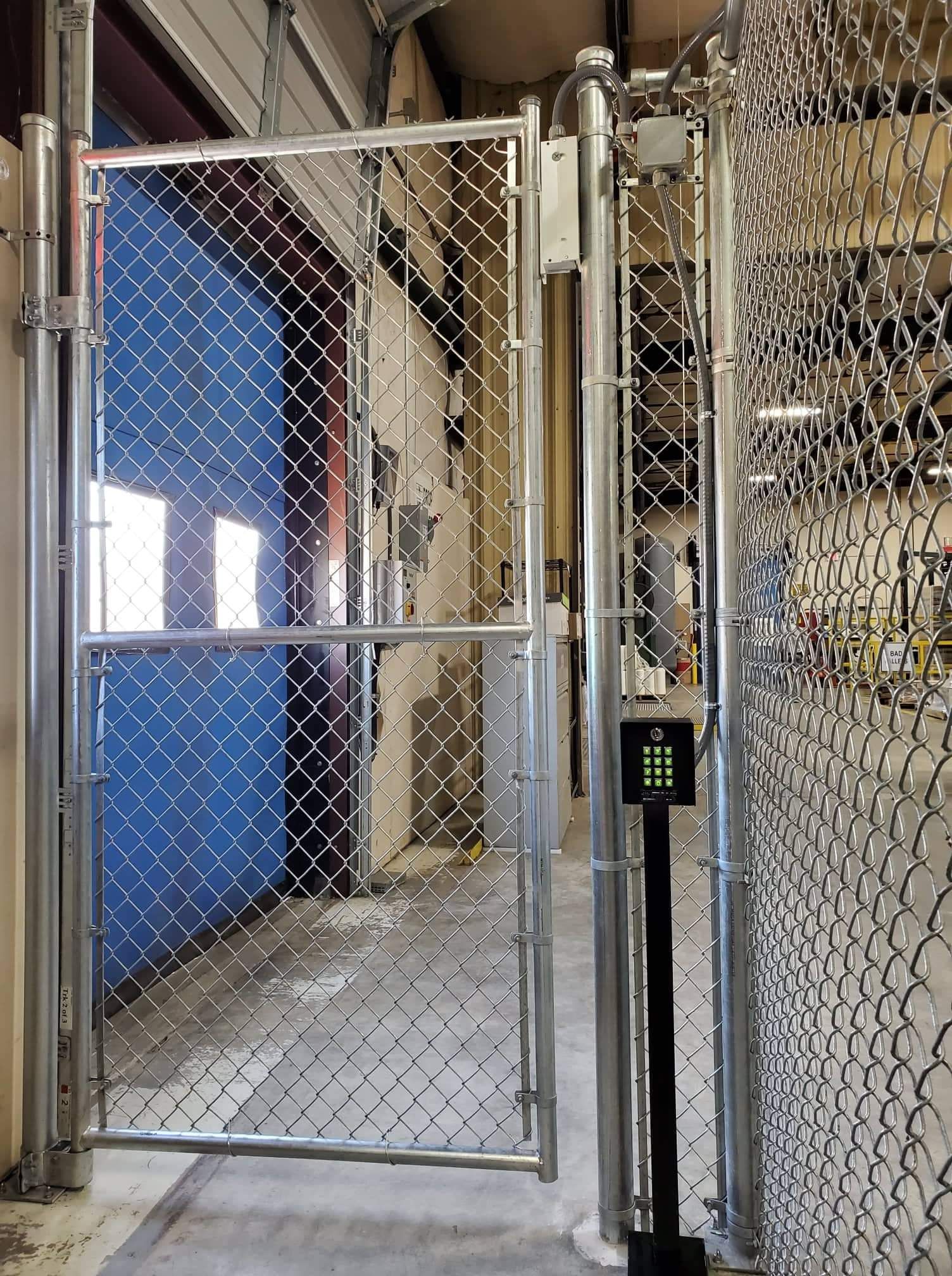 Chain-link gate, open doorway. Code entry pad on right. Gray and blue colors, industrial setting.