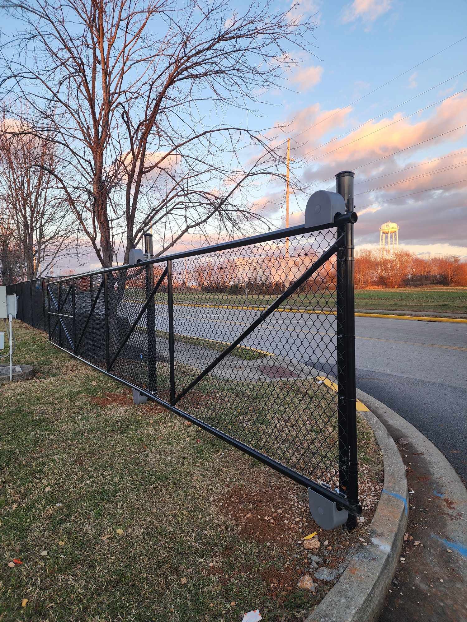 Black chain-link fence along a curved road; evening sky with clouds; brown grass and tree.
