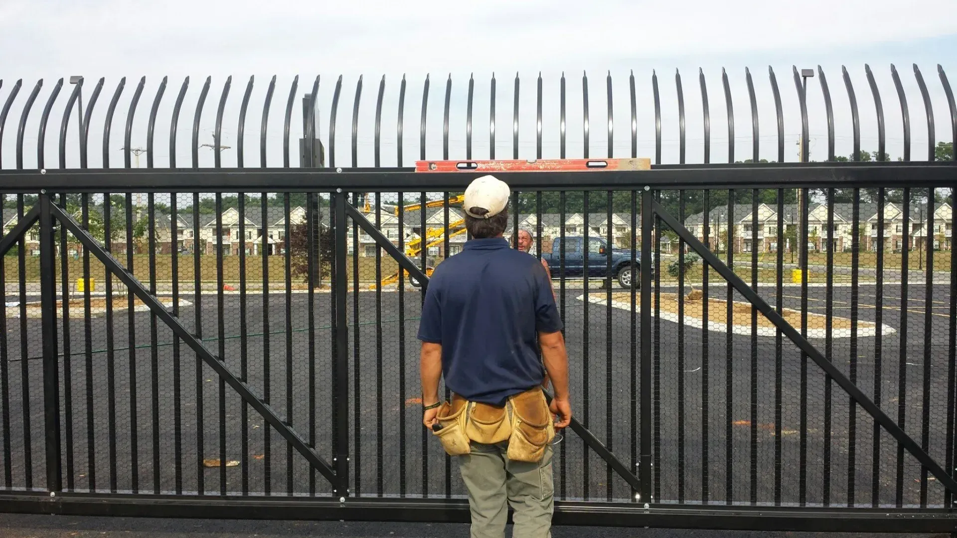 A person, with tools, inspecting a black metal security gate with upward-curving spikes, outdoors.