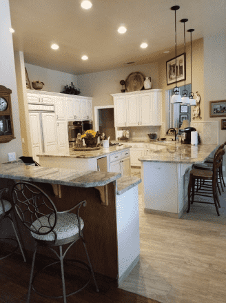 A kitchen with white cabinets , granite counter tops , stools and a clock on the wall.