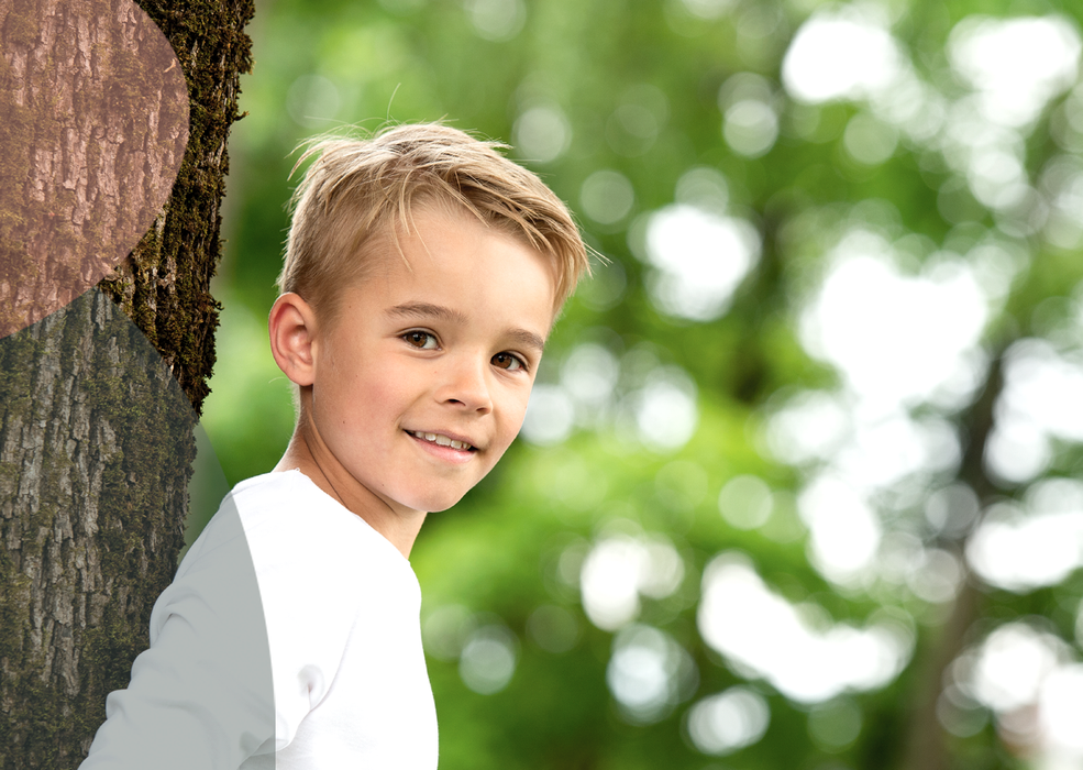 En ung person med lyst hår iført en hvit langermet skjorte lener seg mot en teksturert trestamme i en løvrik park.