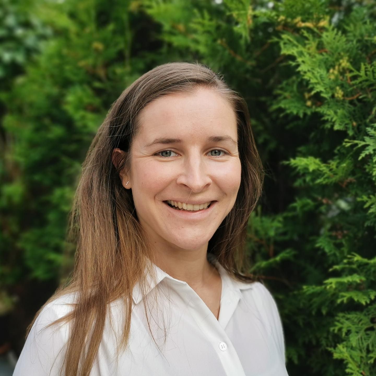 En smilende person med langt brunt hår iført en hvit skjorte med krage, plassert foran grønt, løvrikt løvverk.