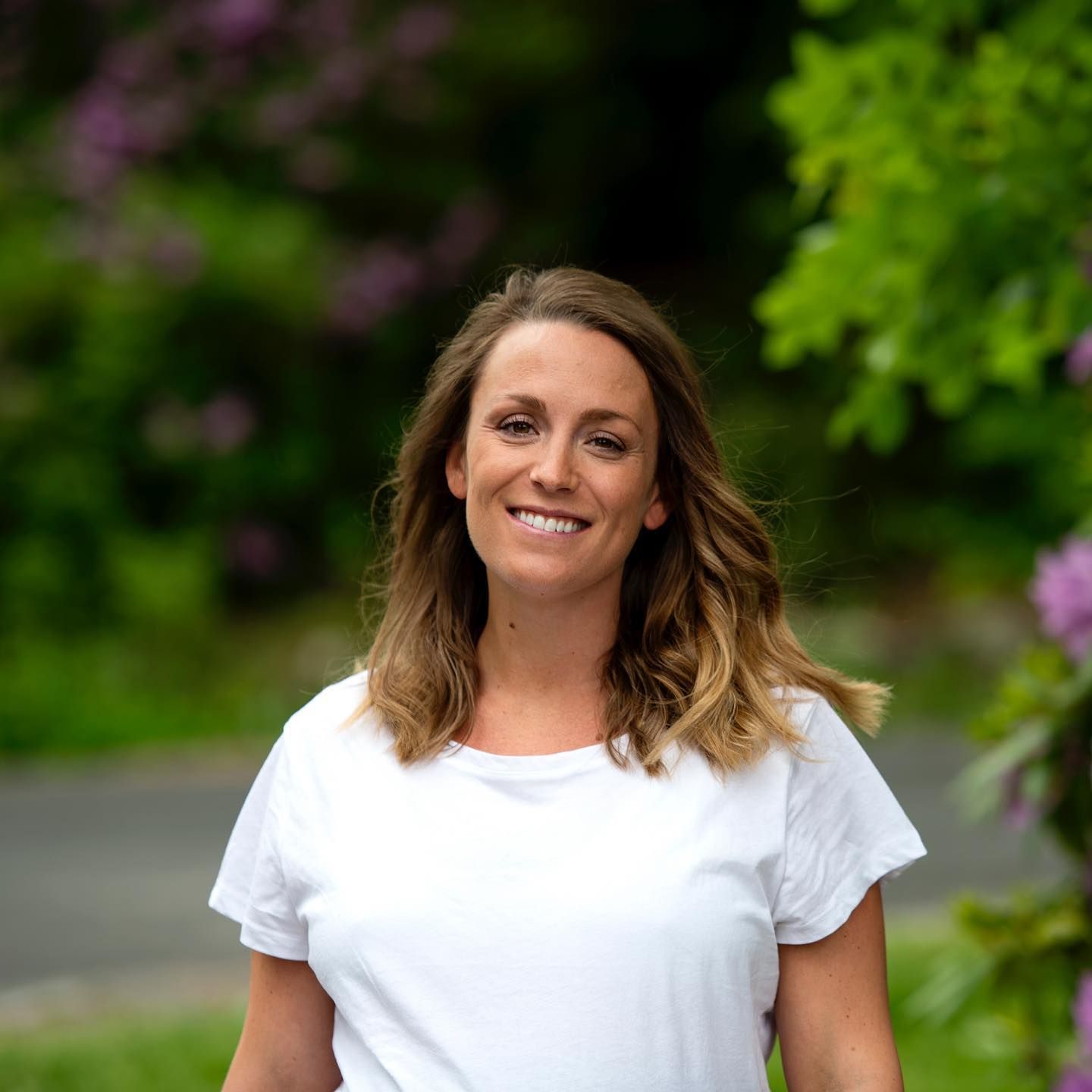 En person med skulderlangt brunt hår iført en hvit t-skjorte, smilende utendørs foran et uskarpt grønt landskap.