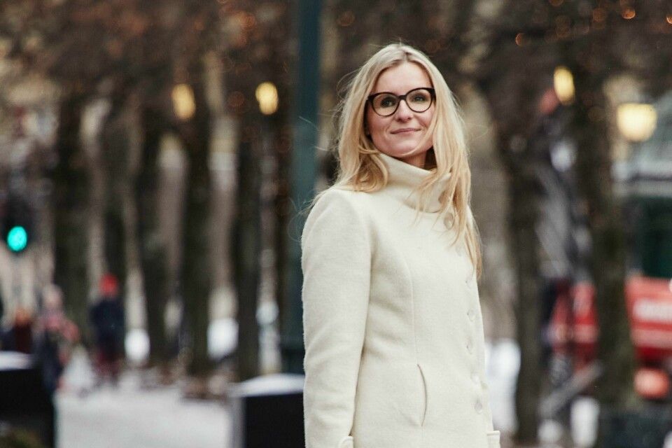 En blond person med briller og kremfarget frakk står utendørs i en trærom om vinteren.