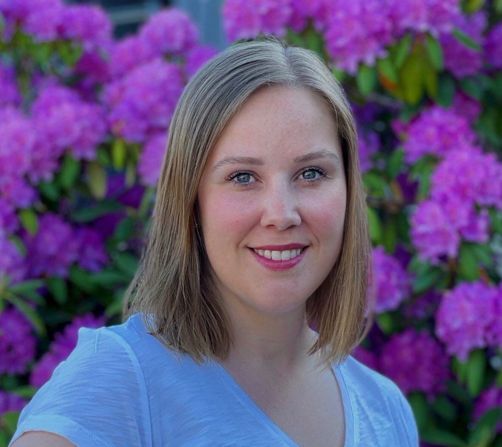 En person med skulderlangt blondt hår smiler foran en busk med livlige lilla rhododendronblomster.