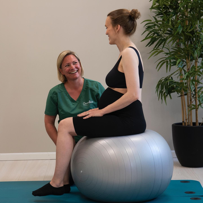 En fysioterapeut i en grønn topp veileder en gravid person som sitter på en sølvfarget treningsball i et studio.