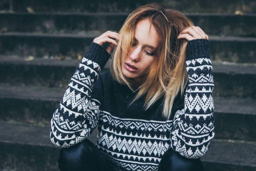 En person med blondt hår iført en mønstret svart-hvitt genser sitter i en betongtrapp med hendene til hodet.