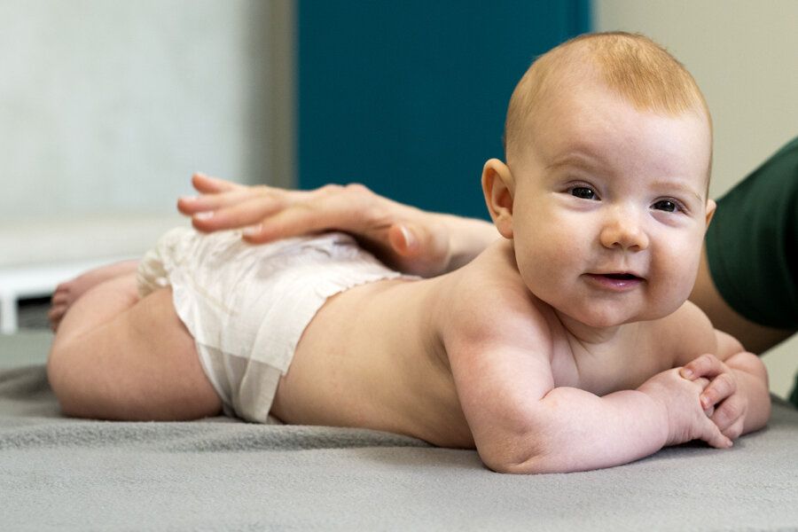 En persons hånd hviler forsiktig på korsryggen til en baby som ligger på magen og ser fremover med et lite smil.