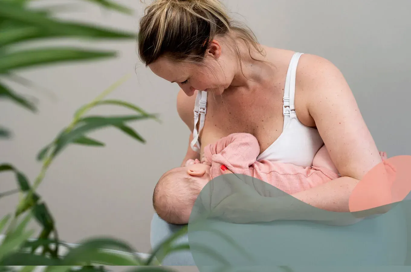 En person i en hvit amme-BH holder og ammer en baby kledd i rosa, med en grønn plante i forgrunnen.