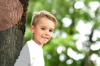 Et smilende barn i hvit skjorte står ved siden av en trestamme med en uskarp grønn bakgrunn.