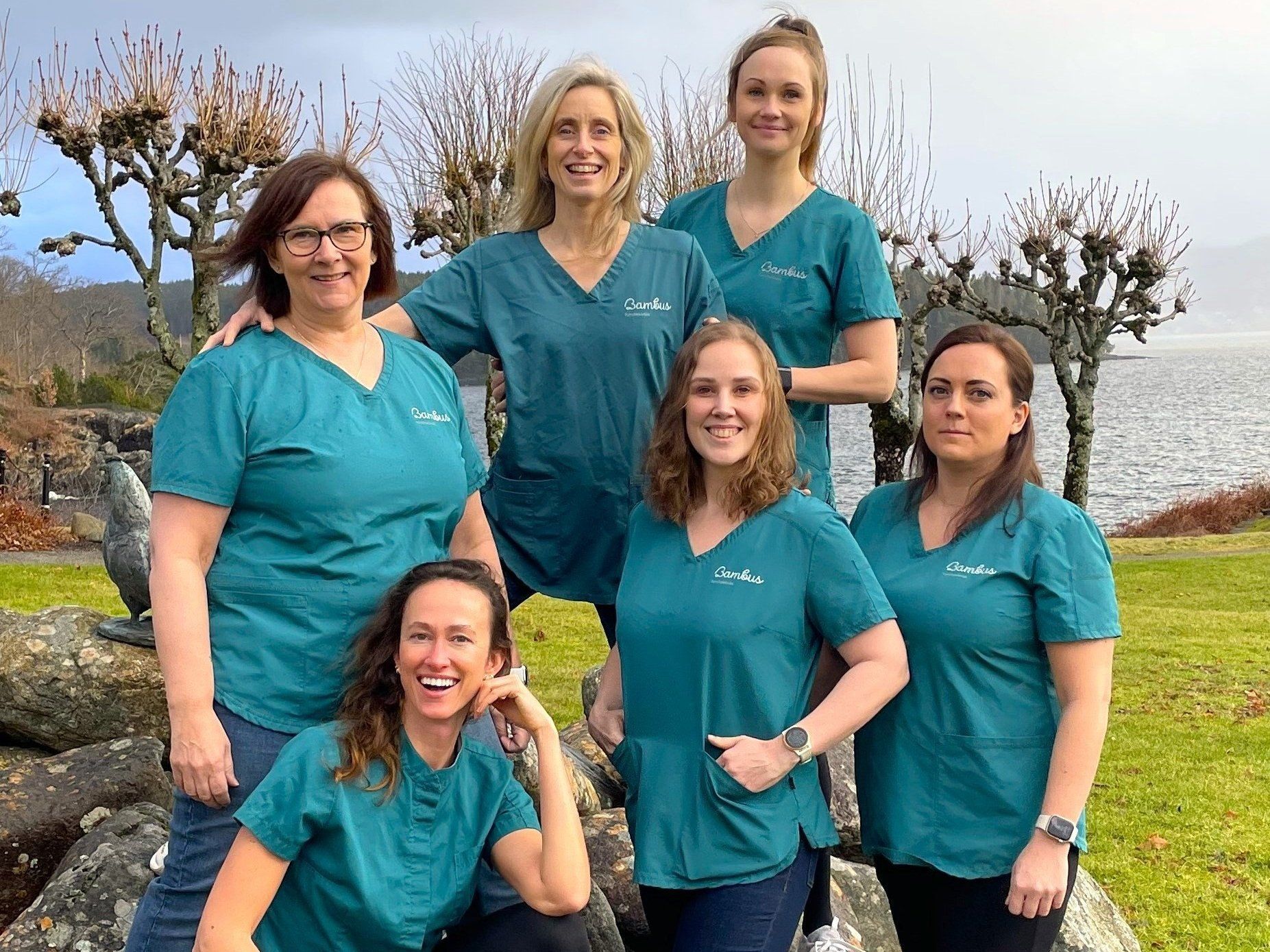 Seks personer i blågrønne skrubber poserer sammen utendørs i nærheten av et vann og trær på en overskyet dag.