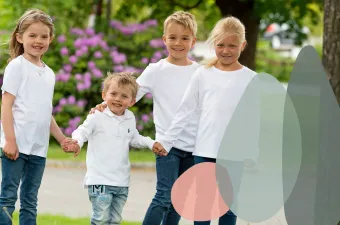 Fire smilende barn kledd i hvite langermede skjorter og jeans som holder hender i en park med lilla blomster.