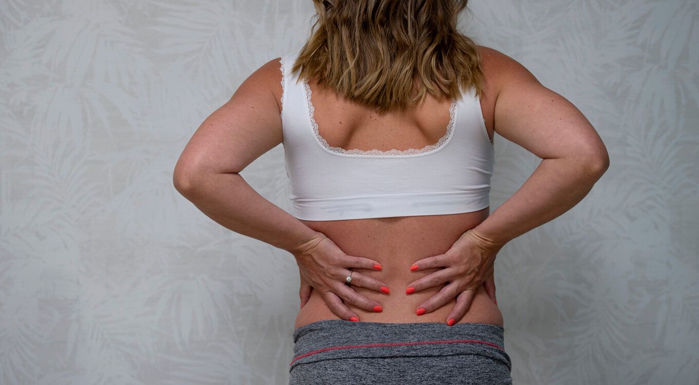 Person som står med ryggen mot kameraet og holder korsryggen med begge hender mot en hvit vegg med tekstur.