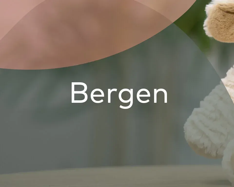 En hvit, luftig teddybjørn sitter på et bord mot en lys bakgrunn med teksten «Bergen» sentrert i hvitt.