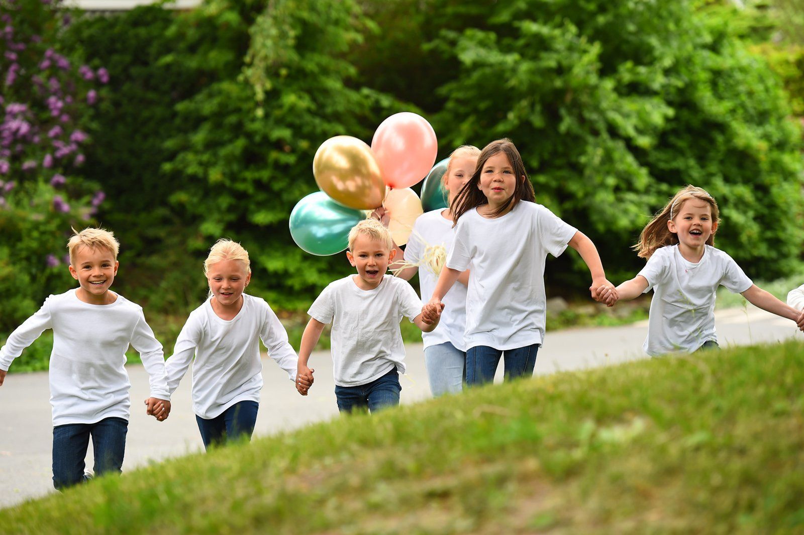 En gruppe barn i hvite skjorter løper sammen utendørs, holder hender og bærer en haug med fargerike ballonger.