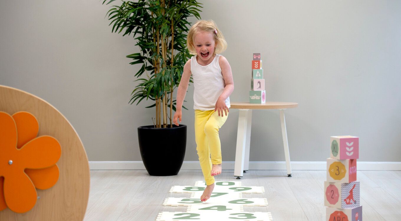 En person i hvit tanktopp og gule bukser hopper på nummererte gulvmatter i et lyst rom med en potteplante.