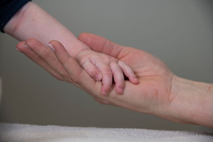 En voksens åpne håndflate støtter forsiktig en liten barnehånd mot en myk, nøytral bakgrunn.