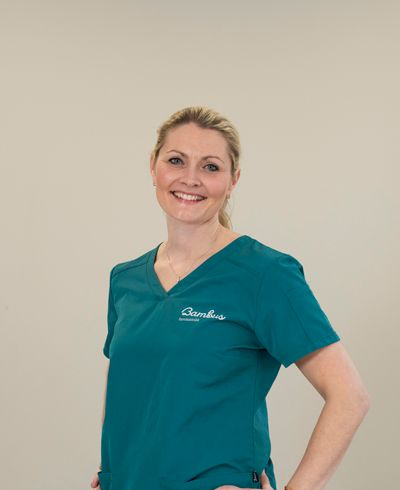 En smilende profesjonell iført en blågrønn V-hals skrubbtopp med brodert logo, mot en ensfarget beige bakgrunn.