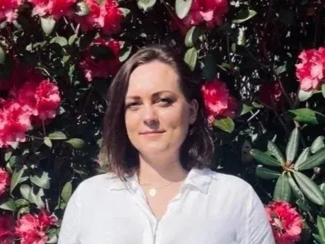 En person med skulderlangt mørkt hår iført hvit skjorte, poserer foran knallrosa blomstrende rododendroner.