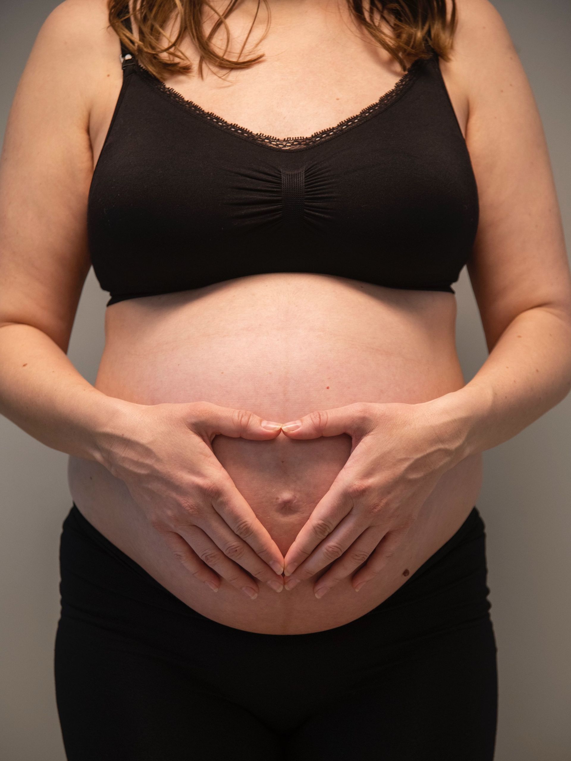 En gravid person iført svart BH og bukser former en hjerteform med hendene over magen.