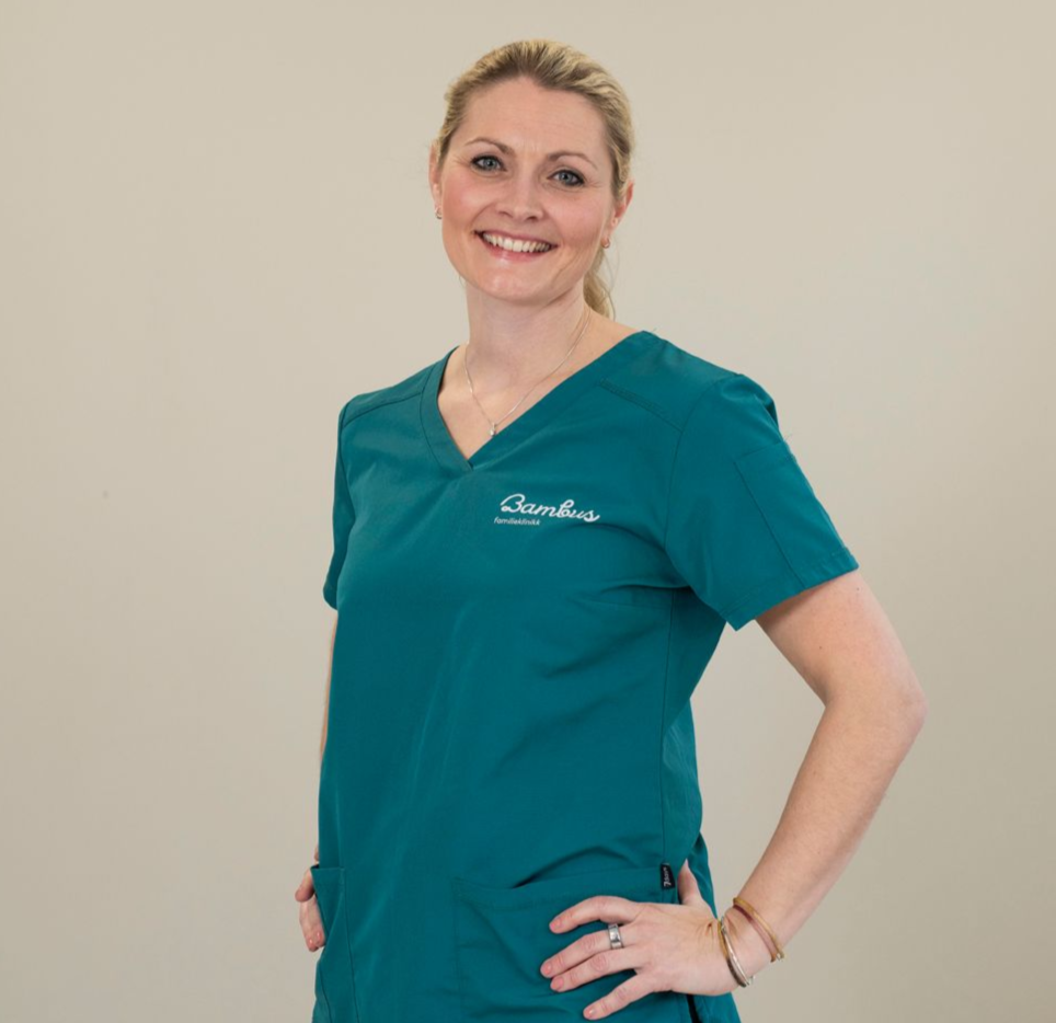 En smilende profesjonell, stående med hendene i siden, iført en mørk blågrønn skrubbtopp med en liten hvit logo på brystet.