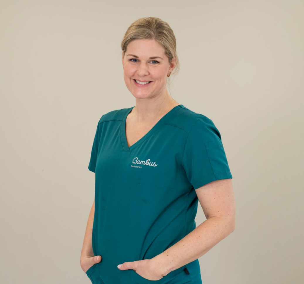 En smilende person, iført en blågrønn skrubbtopp med logo, står med hendene i lommene mot en enkel lys bakgrunn.