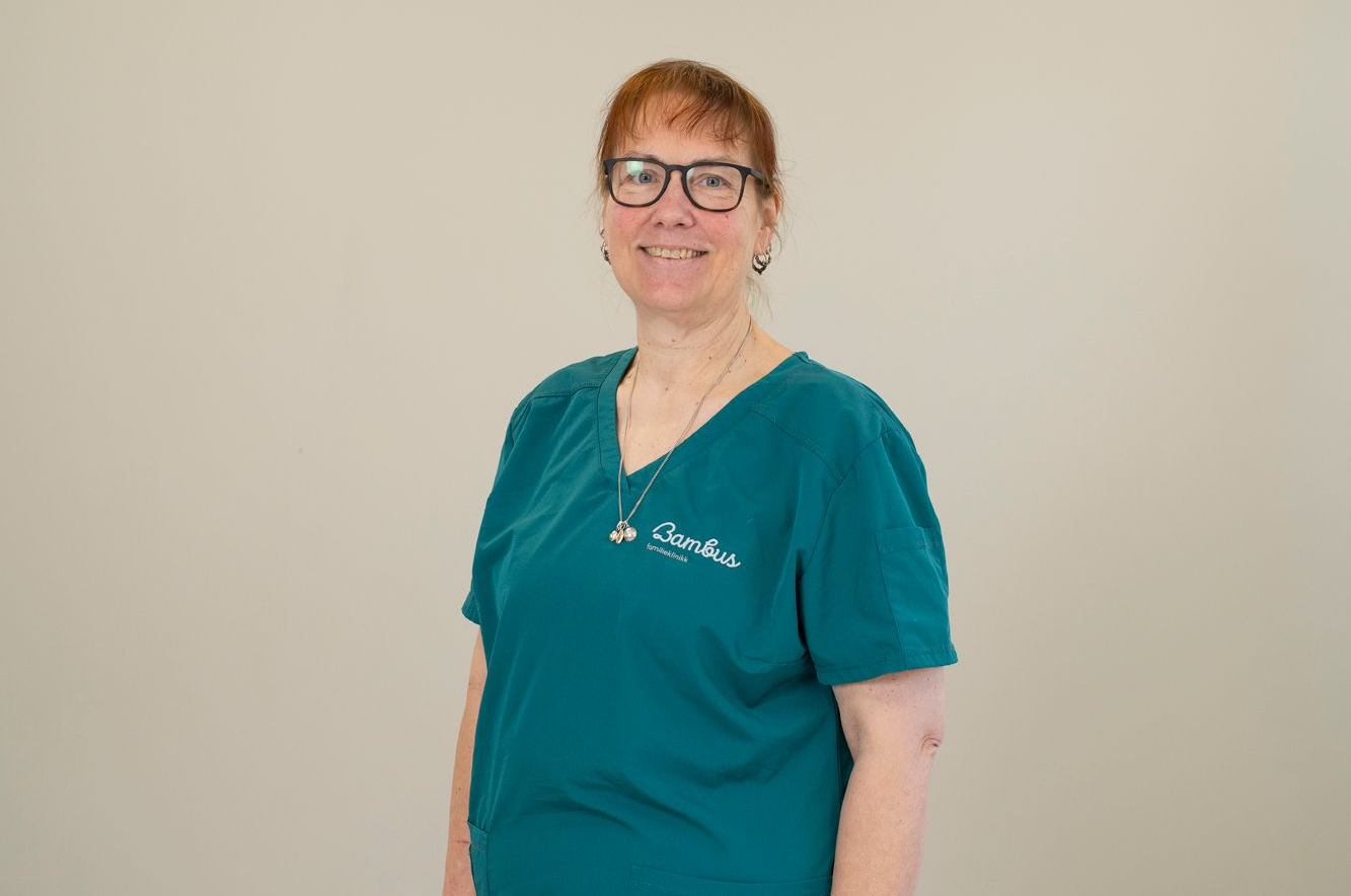 En person med briller og en blågrønn skrubbtopp med logo, smilende mot en enkel lys bakgrunn.