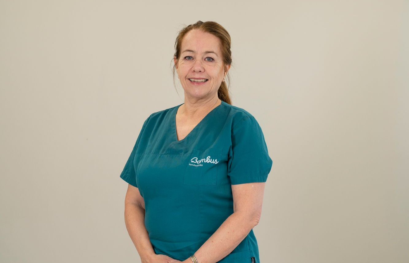 En person iført en blågrønn V-hals skrubbtopp med brodert logo, stående mot en nøytral bakgrunn.