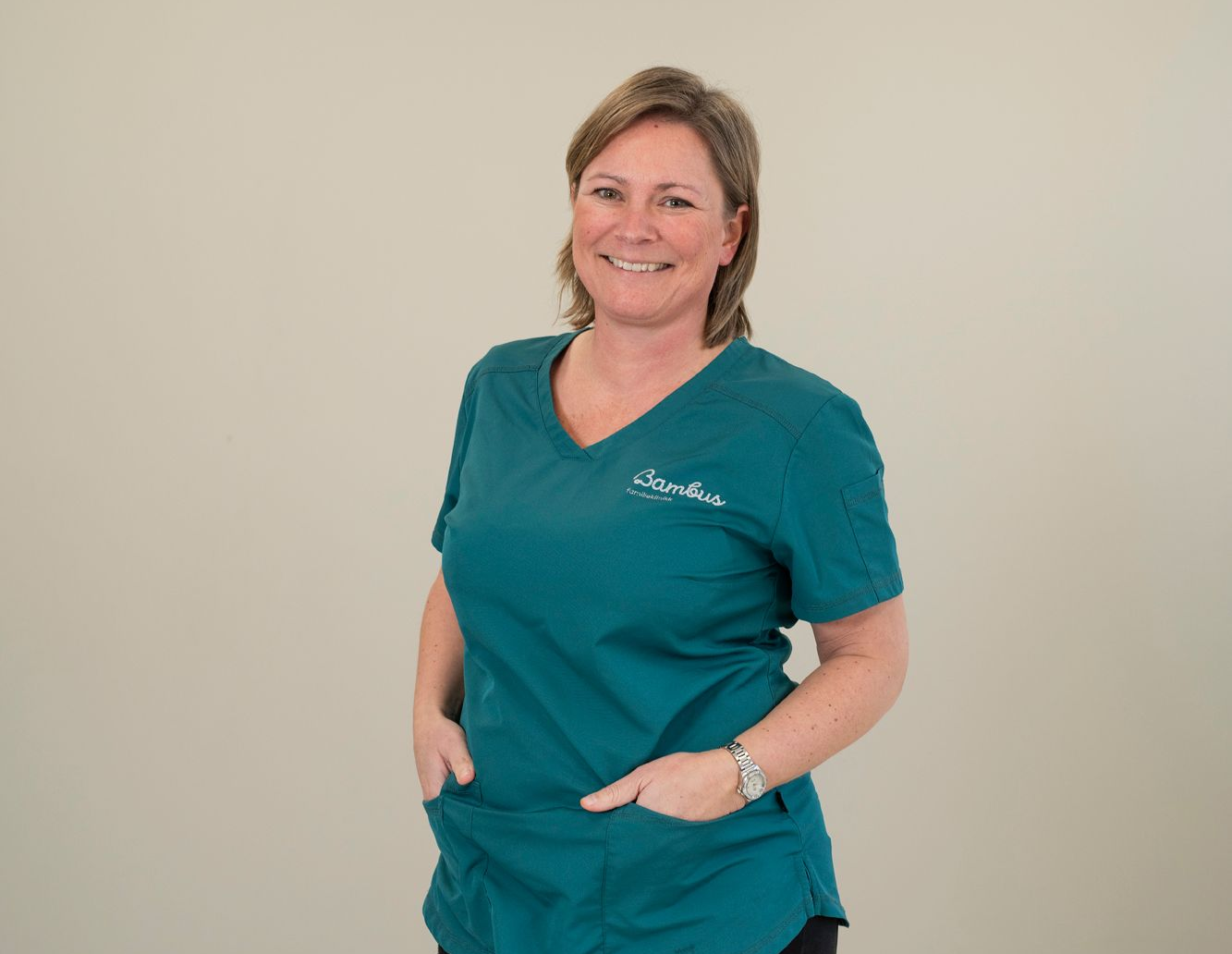 En smilende person i en blågrønn skrubbtopp med hendene stukket i lommene foran, posert mot en enkel bakgrunn.